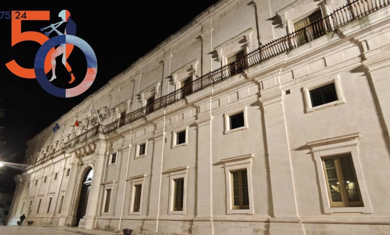 Vista notturna del Palazzo Ducale di Martina Franca illuminato, con il logo del 50° Festival della Valle d'Itria visibile in primo piano. Una facciata elegante e bianca con archi e balconi in ferro battuto caratterizza questo storico edificio. Le bandiere dell'Unione Europea e dell'Italia ondeggiano sul balcone principale. Foto di Anton Pio Rubino.