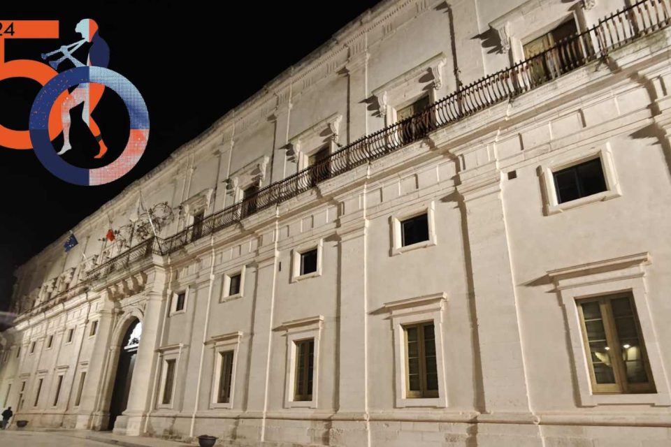 Vista notturna del Palazzo Ducale di Martina Franca illuminato, con il logo del 50° Festival della Valle d'Itria visibile in primo piano. Una facciata elegante e bianca con archi e balconi in ferro battuto caratterizza questo storico edificio. Le bandiere dell'Unione Europea e dell'Italia ondeggiano sul balcone principale. Foto di Anton Pio Rubino.