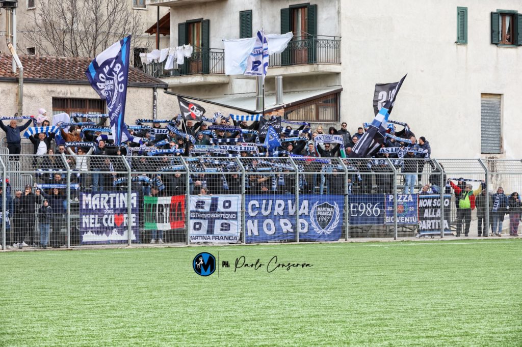 Curva Nord del Martina Calcio con tifoseria organizzata in trasferta, bandiere e striscioni esposti durante una gara di campionato.